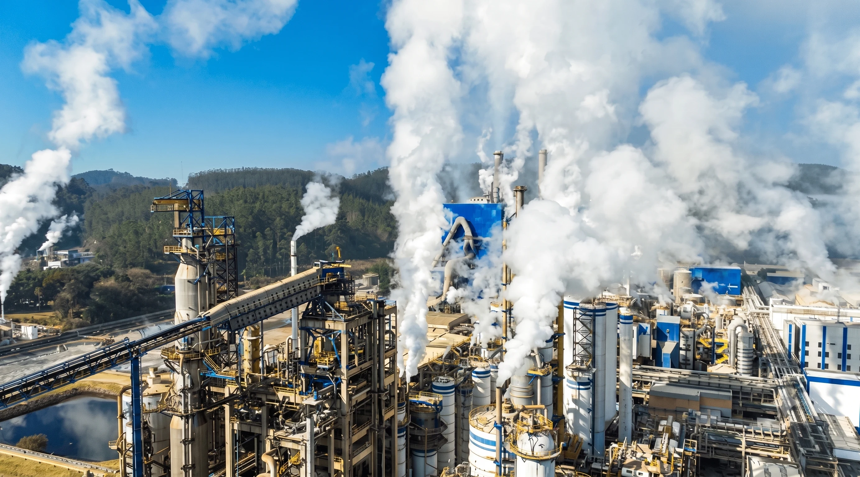 Vista aérea de un complejo industrial con columnas de vapor, correas transportadoras y cerros boscosos al fondo.